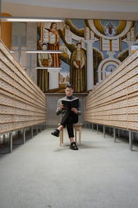A focused young student reading in a library between rows of drawers with a mural in the background.