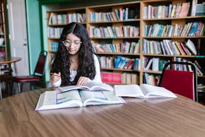 A young woman studying at a library table surrounded by bookshelves, focused on her work.